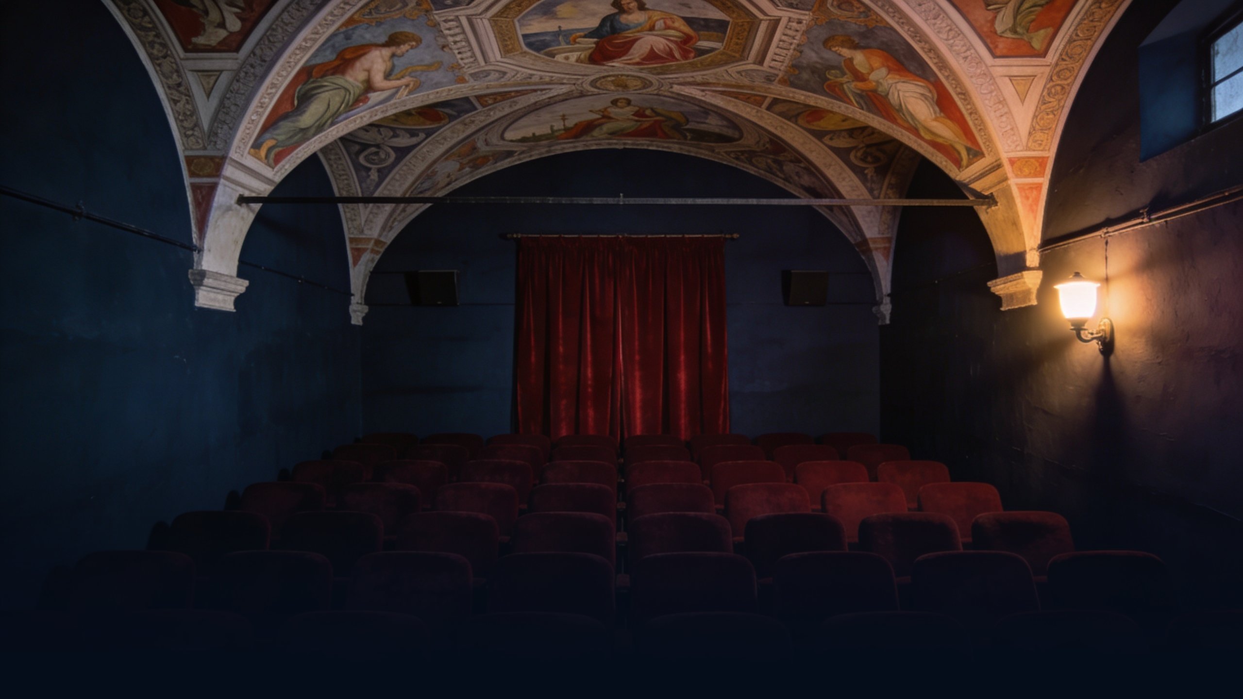 Empty historic Italian cinema interior at dusk with frescoed vaulted ceiling, rows of empty deep red velvet seats facing a closed red velvet curtain, single dim wall sconce providing warm tungsten light