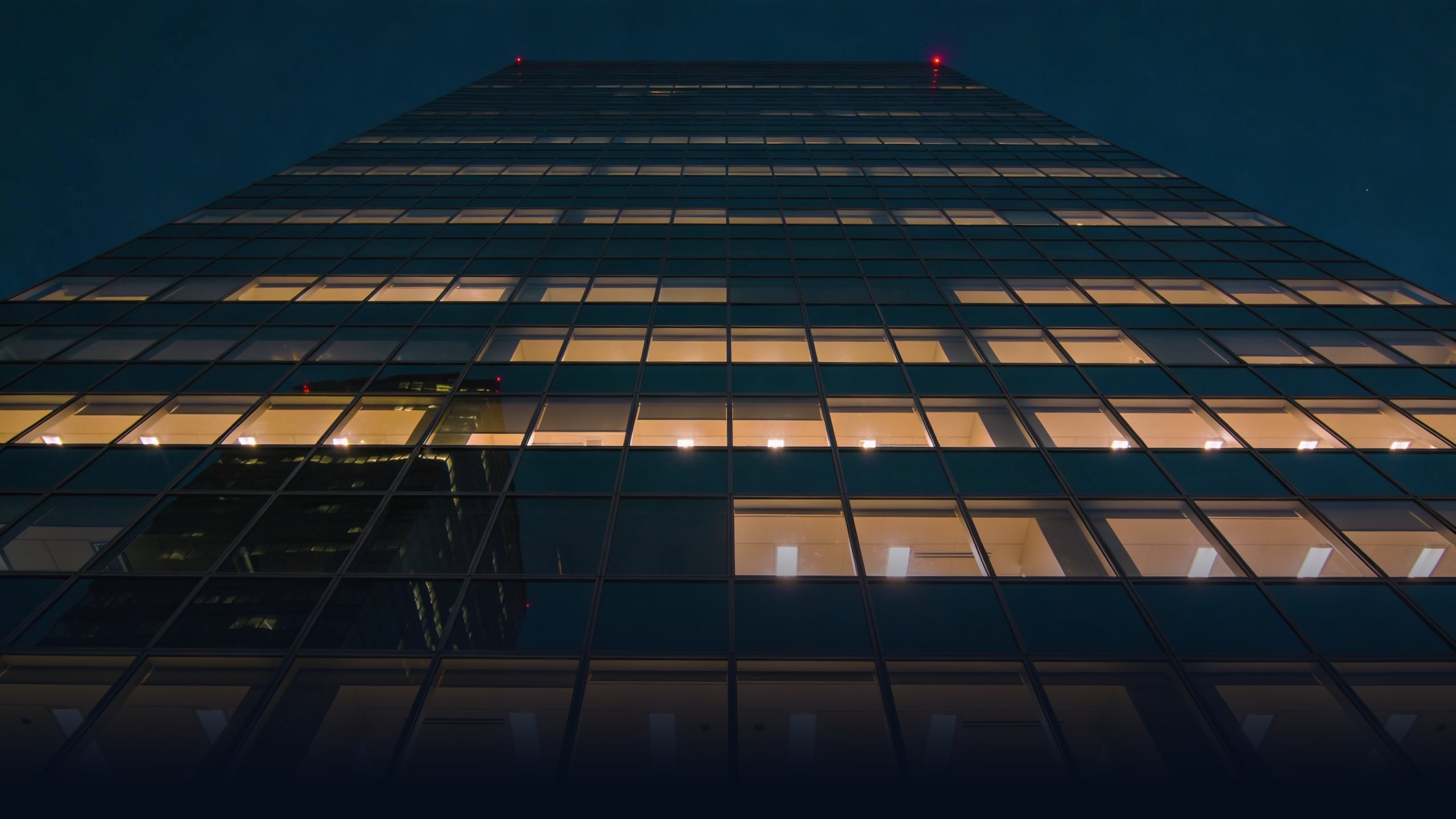 Modern Los Angeles corporate office tower at night, mirrored glass facade with backlit windows in warm tungsten light against a deep navy sky
