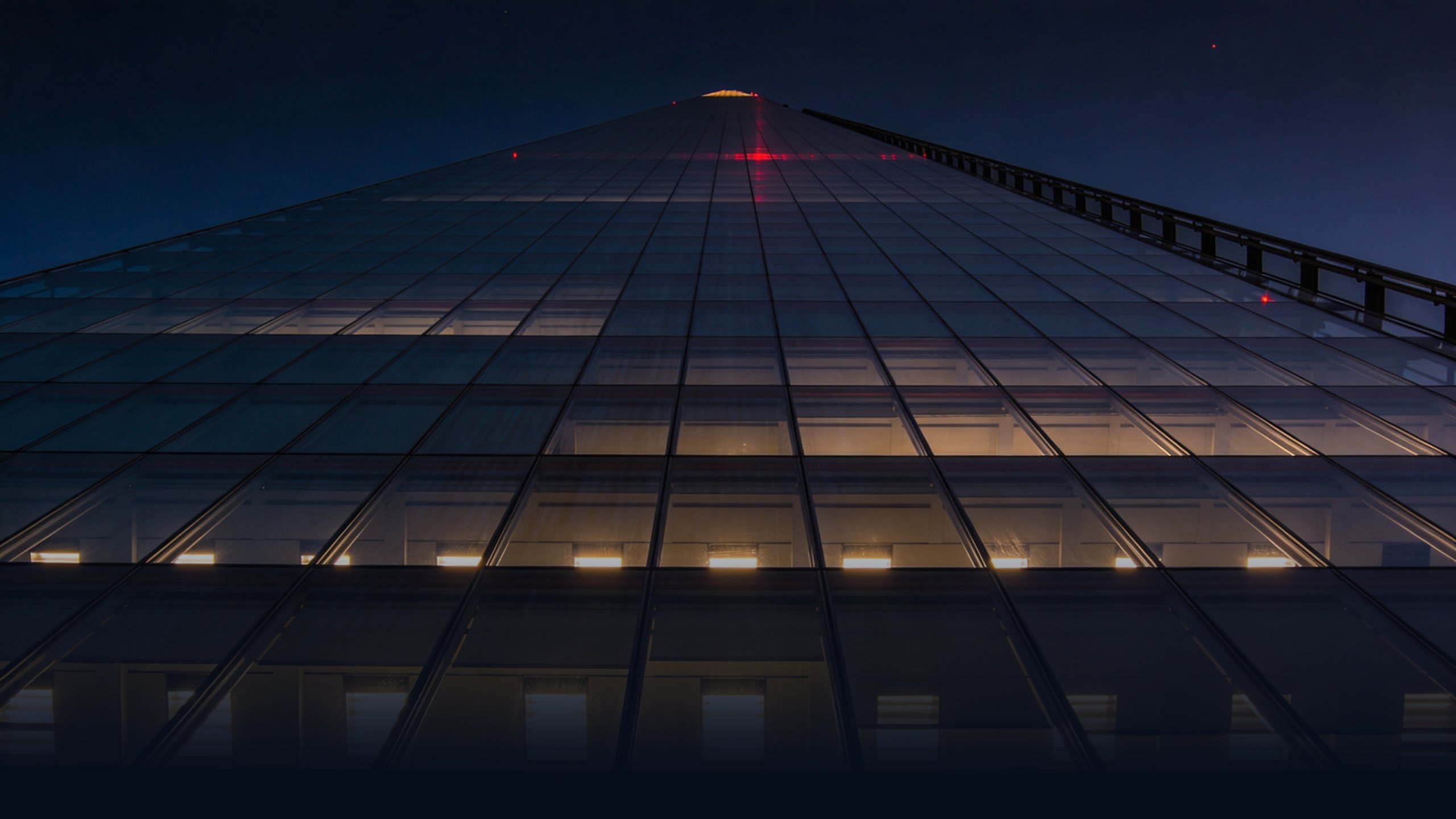 Modern London corporate glass office tower at night photographed from a low ground-level angle with backlit windows glowing warm tungsten against deep navy sky