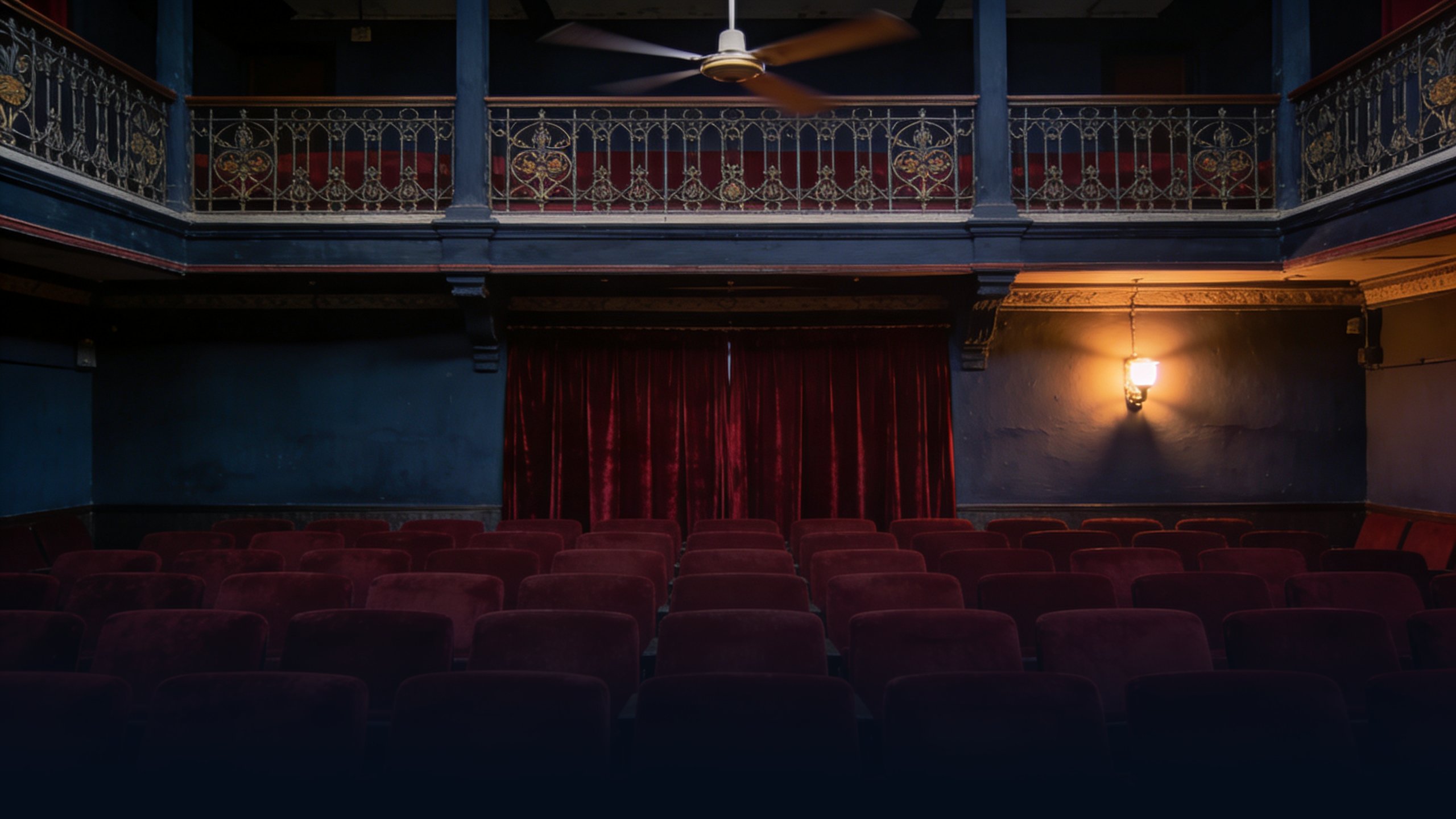 Empty historic Indian single-screen cinema interior at dusk, ornate balcony with wrought-iron railings, rows of empty deep red velvet seats, single overhead ceiling fan captured with slight motion blur