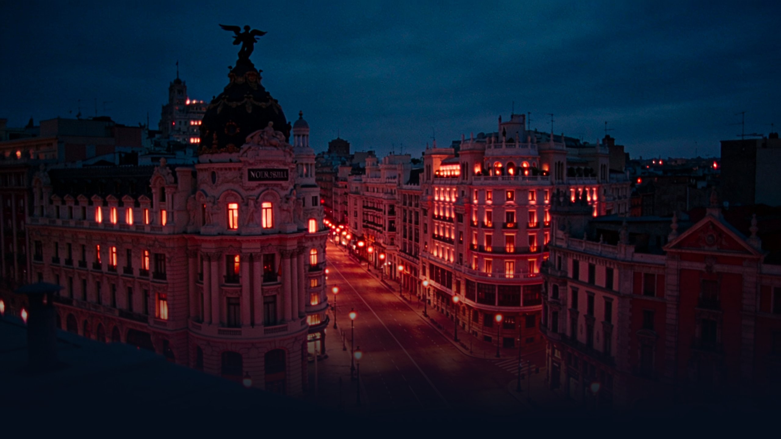 Madrid Gran Vía photographed from a rooftop at blue hour, iconic curved Beaux-Arts buildings lining the avenue with warm tungsten interior lights against deep navy twilight sky, stone domes silhouetted overhead