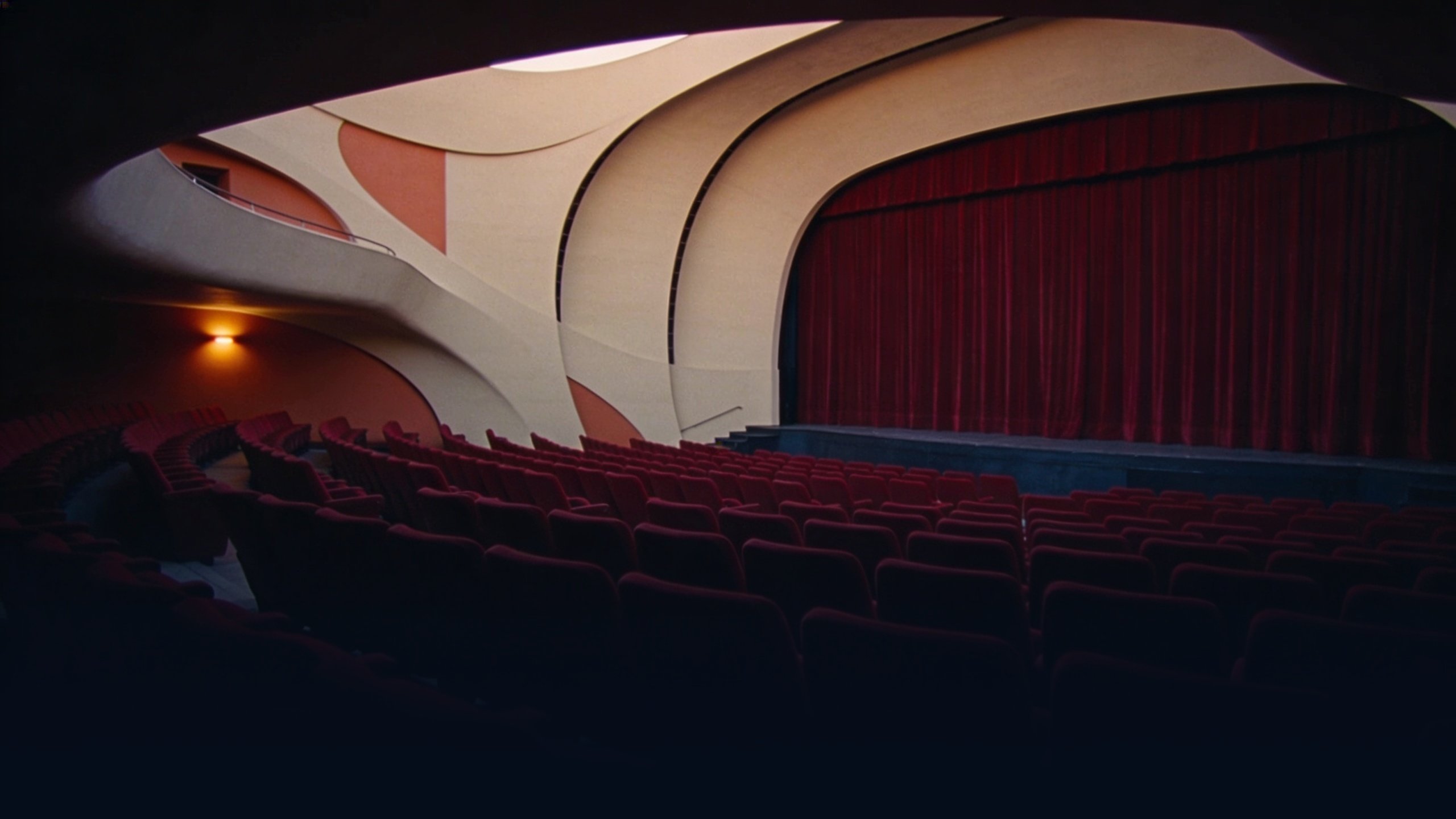 Empty Brazilian modernist 1950s cinema interior at dusk with curved organic Niemeyer-style walls in warm beige and terracotta tones, rows of empty seats in deep red, single low ambient amber light source