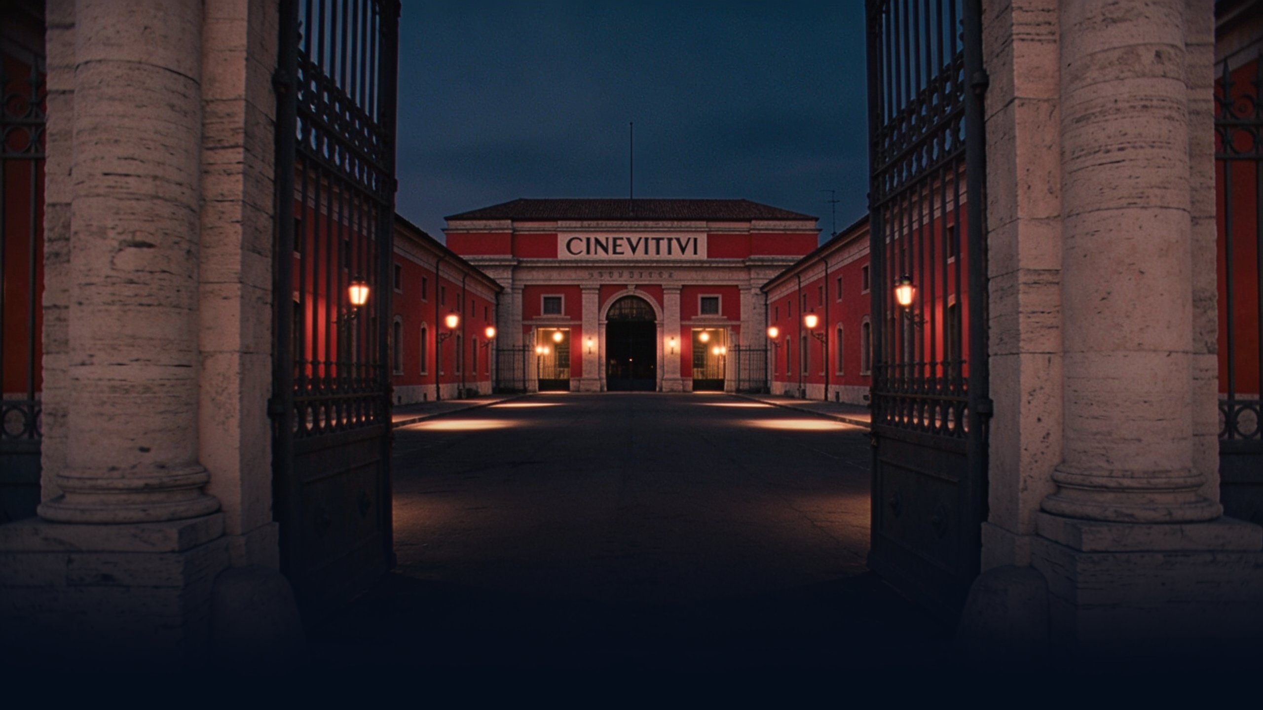 Cinecittà studio gates at dusk photographed from outside looking through the wrought-iron entrance, stone gate columns framing the empty avenue beyond with warm tungsten lamps inside the studio grounds