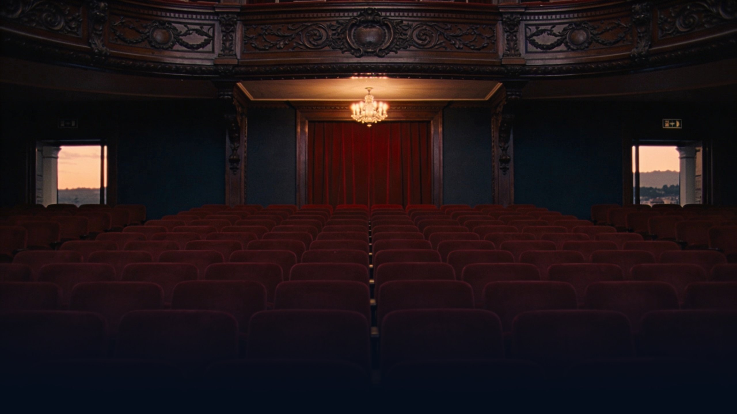 Empty Spanish historic theater interior at dusk with ornate carved wooden balcony fronts in dark mahogany, rows of empty deep red velvet seats, single chandelier above casting warm amber tungsten light