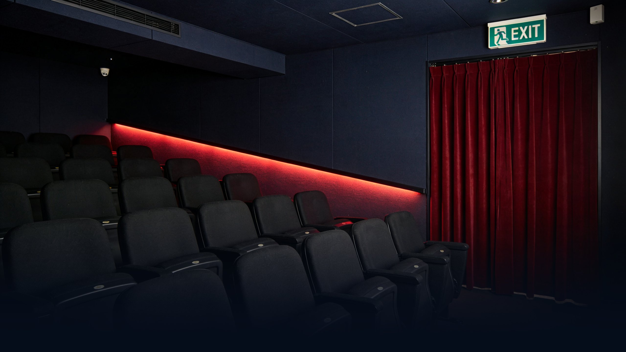Empty small Tokyo independent cinema interior at dusk with tight minimalist auditorium, rows of empty black fabric seats facing a closed deep red velvet curtain, single low-mounted ambient red light strip along the side walls